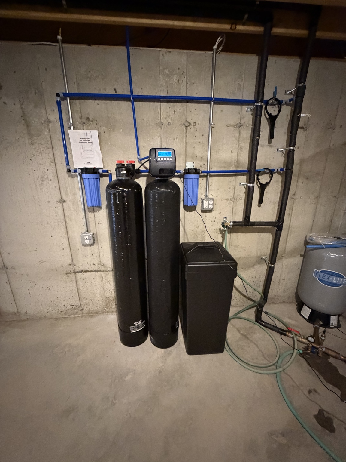 Water softener, whole-house carbon tank, and brine tank with two blue 10-inch filter housings against a concrete basement wall.