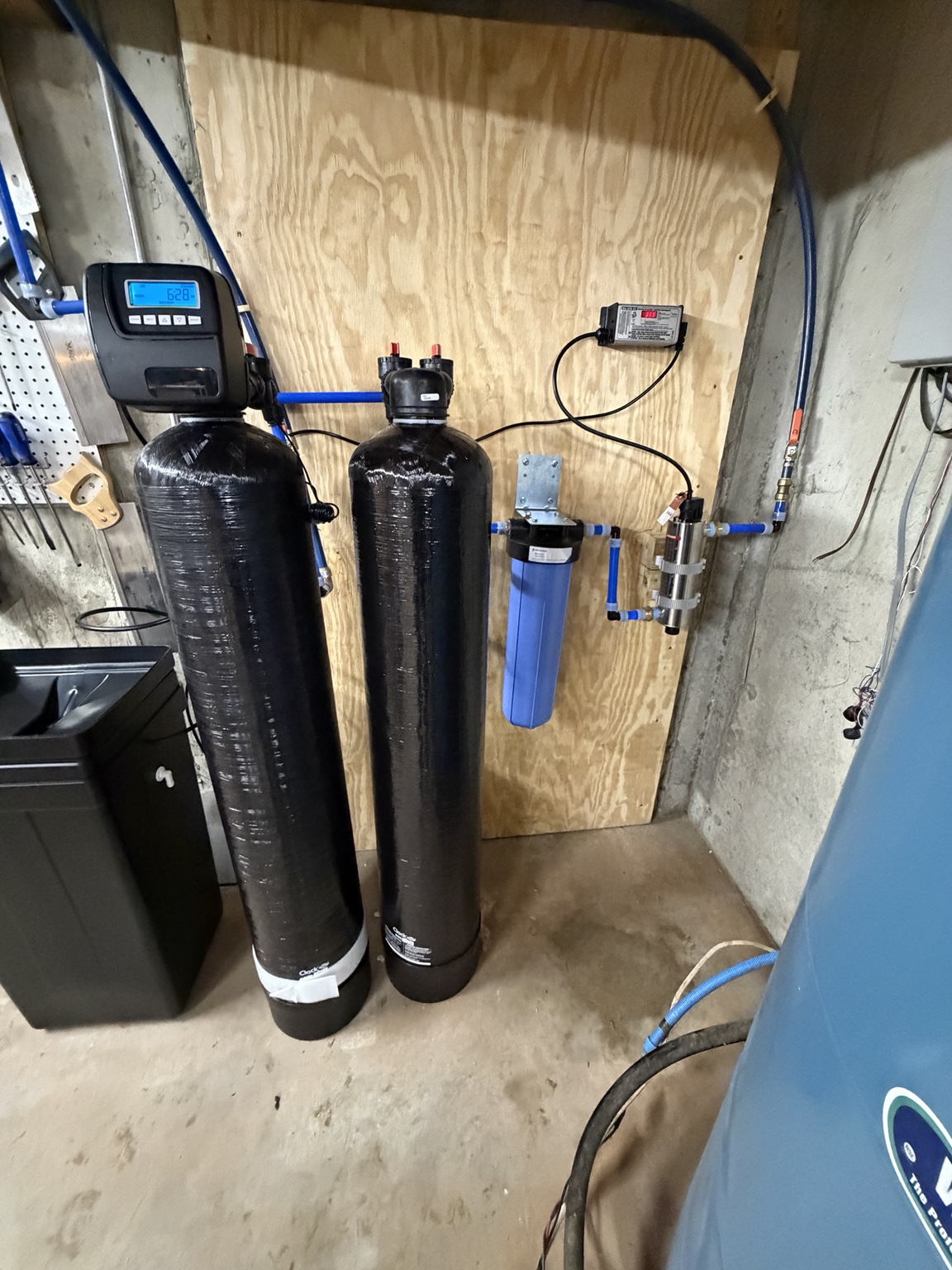 Water softener and whole-house carbon tank beside a 20-inch Big Blue carbon block filter and stainless-steel UV unit mounted on plywood.